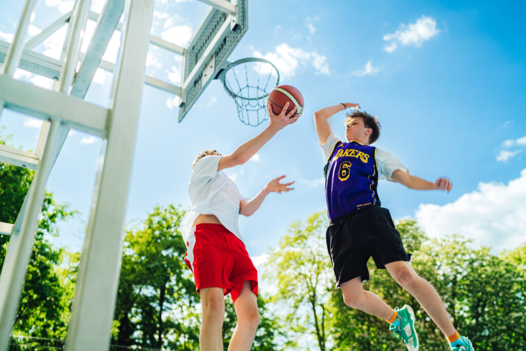 zwei Jungen spielen Basketball, links ein Korb, in den einer einen Ball wirft, der andere blockt ihn