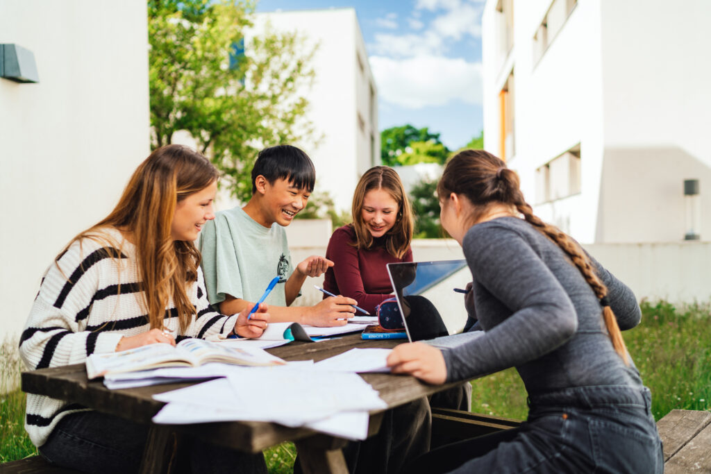 drei Mädchen, ein Junge sitzen draußen an einem Tisch, auf dem Tisch liegen Bücher und Papier, ein Laptop, sie lachen